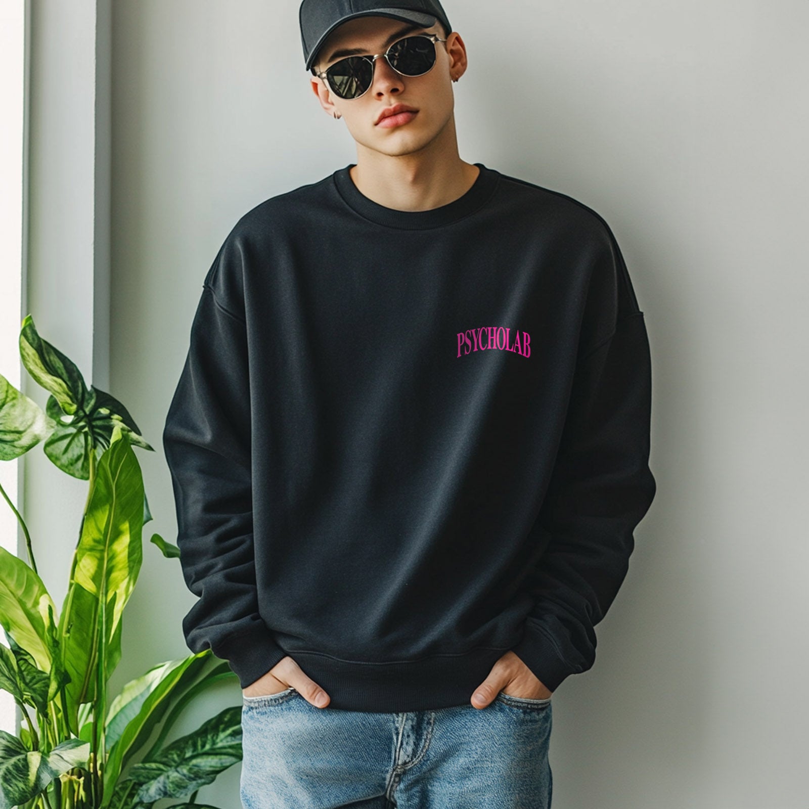Ein Modell trägt das Sweatshirt Knowledge Black von PsychoLab, ein schwarzes Sweatshirt mit auffälligem Logo auf der Brust, während es lässig gegen eine helle Wand lehnt, umgeben von Pflanzen.